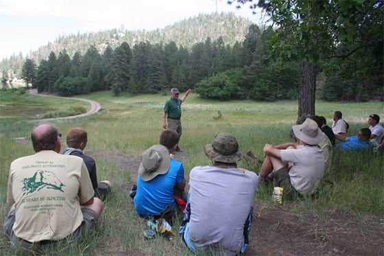 a teacher talking to a crowd of students in a field surrounded by trees and a mountain